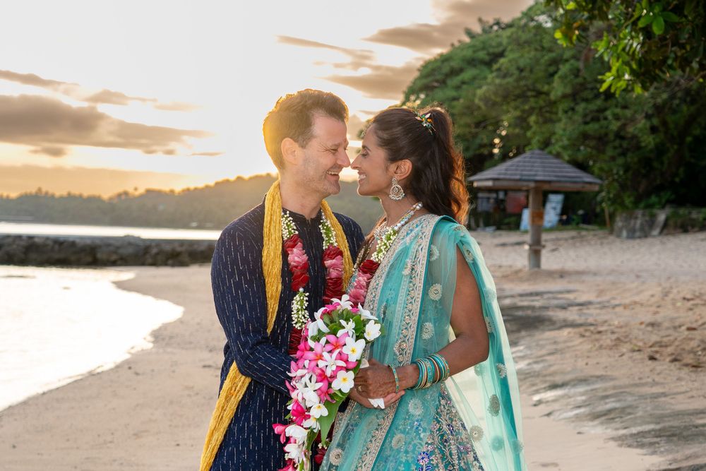 A couple in traditional wedding attire share a romantic moment on a beach at sunset with pink tropical flowers and ocean views.