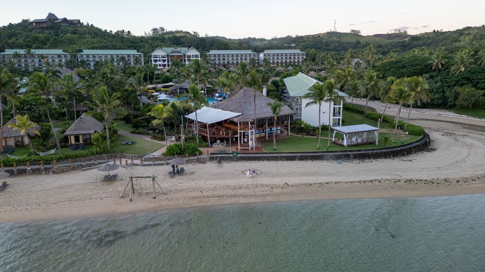 Aerial view of a beachfront resort with palm trees and modern buildings along a curved shoreline.