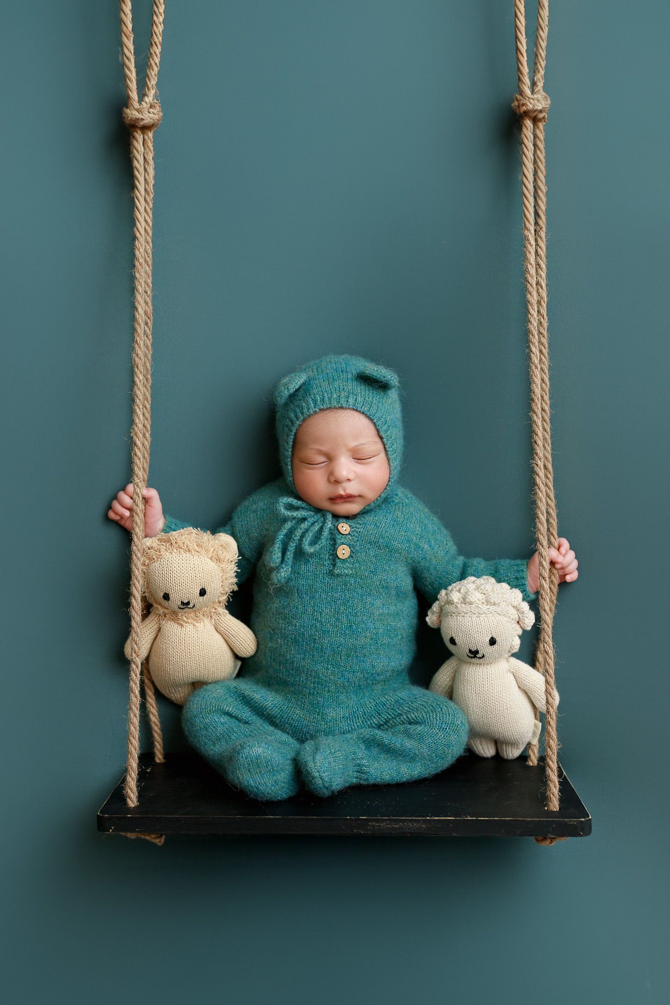 Small figure in teal knitwear rests on wooden swing with two cream-colored teddy bears against teal background in cedar park newborn studio