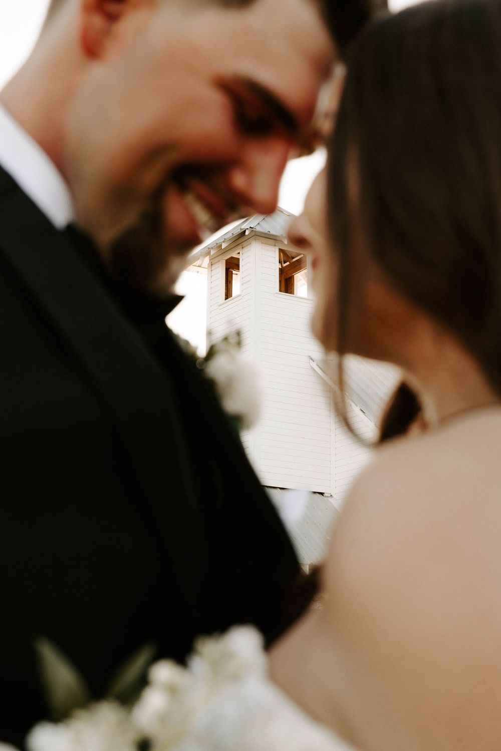 A romantic close-up wedding moment captured in soft focus with a white building visible in the background.