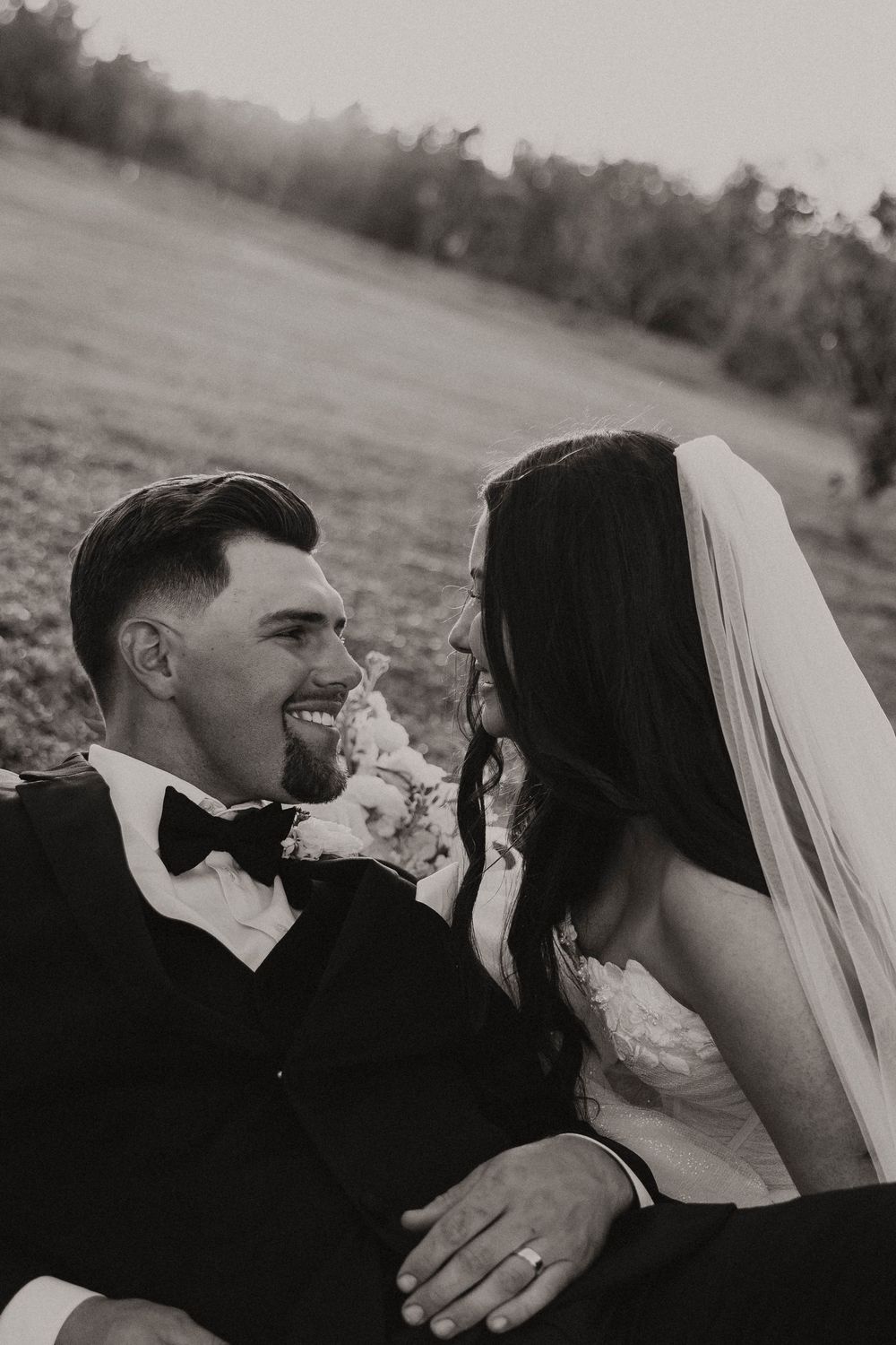 A black and white wedding photo capturing a candid moment between a couple sharing a laugh in an outdoor setting.