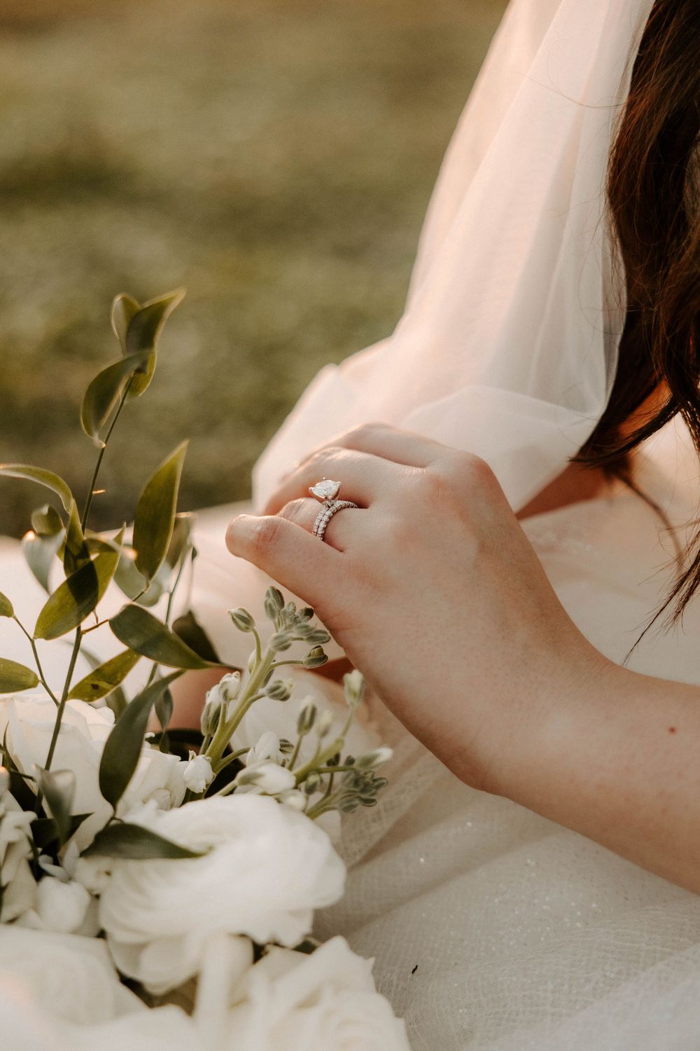 Close up of a bride's hand with engagement ring holding a delicate white floral bouquet with greenery in soft lighting.