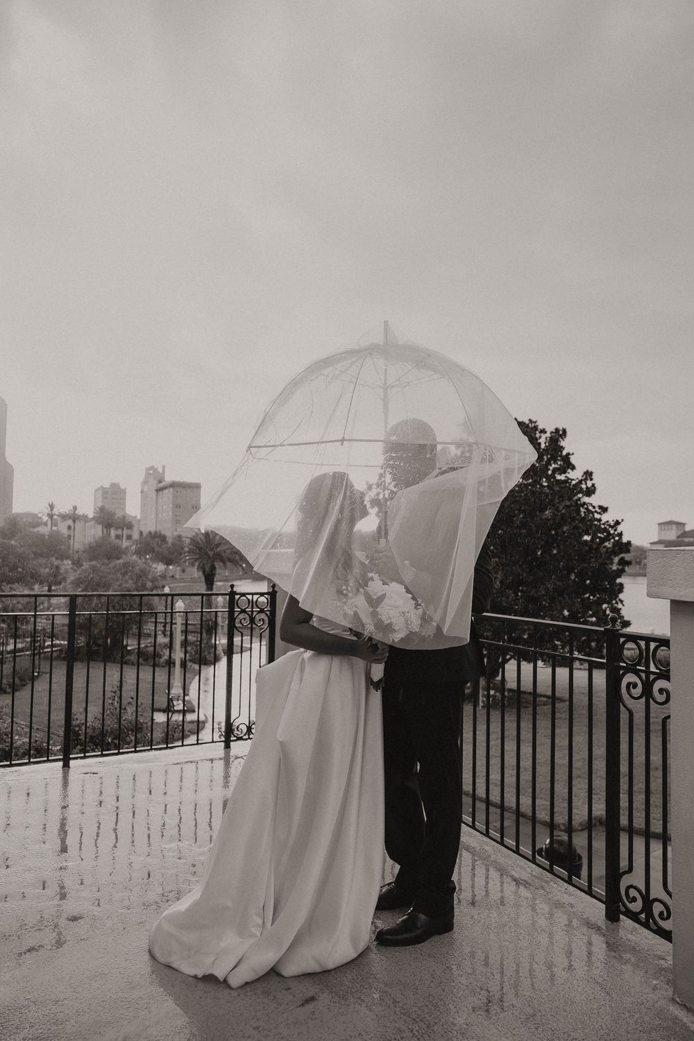 Bride and groom share a romantic moment under a clear umbrella on a rainy wedding day on an outdoor balcony.
