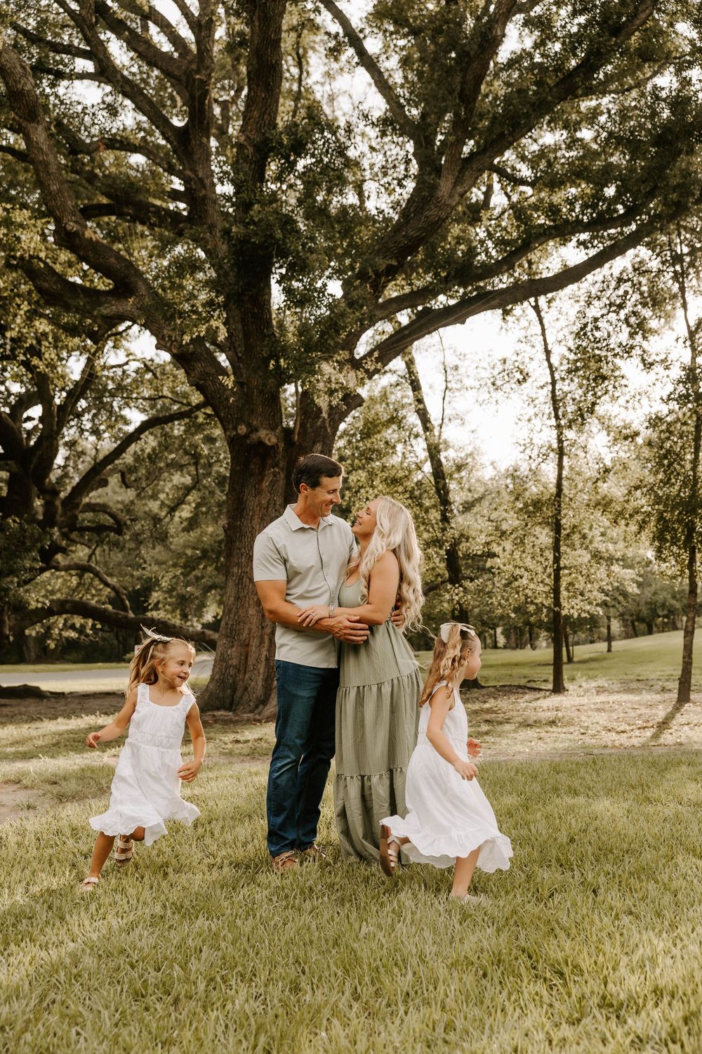 A happy family of four poses outdoors under a large oak tree, with two young girls in white dresses spinning around.