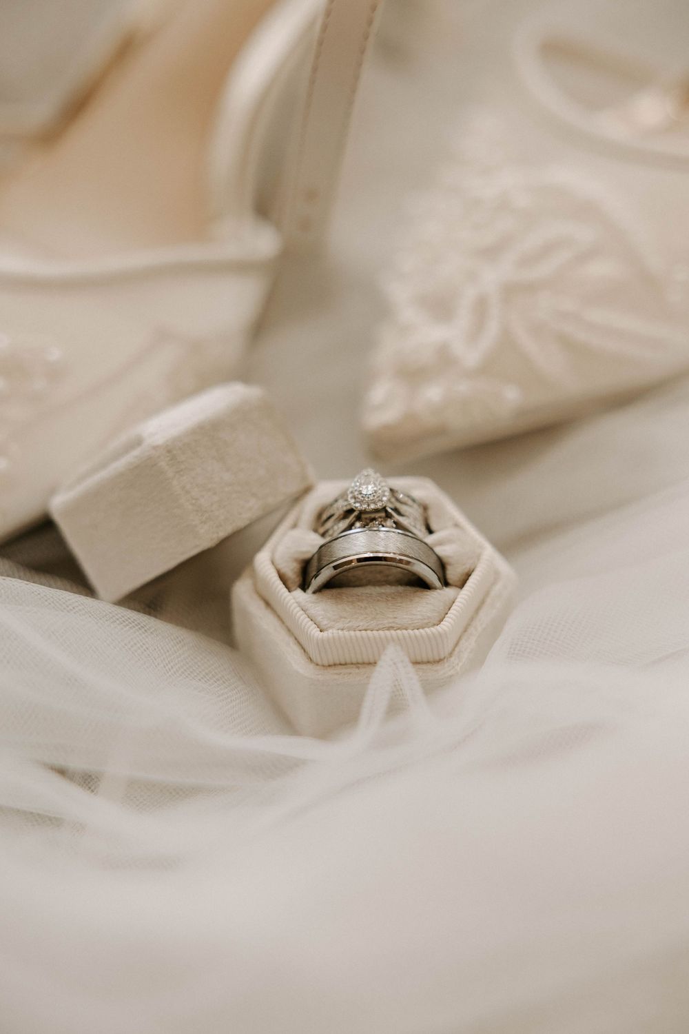 Wedding rings displayed in a hexagonal ivory velvet ring box beside elegant white lace bridal heels on sheer veil fabric.