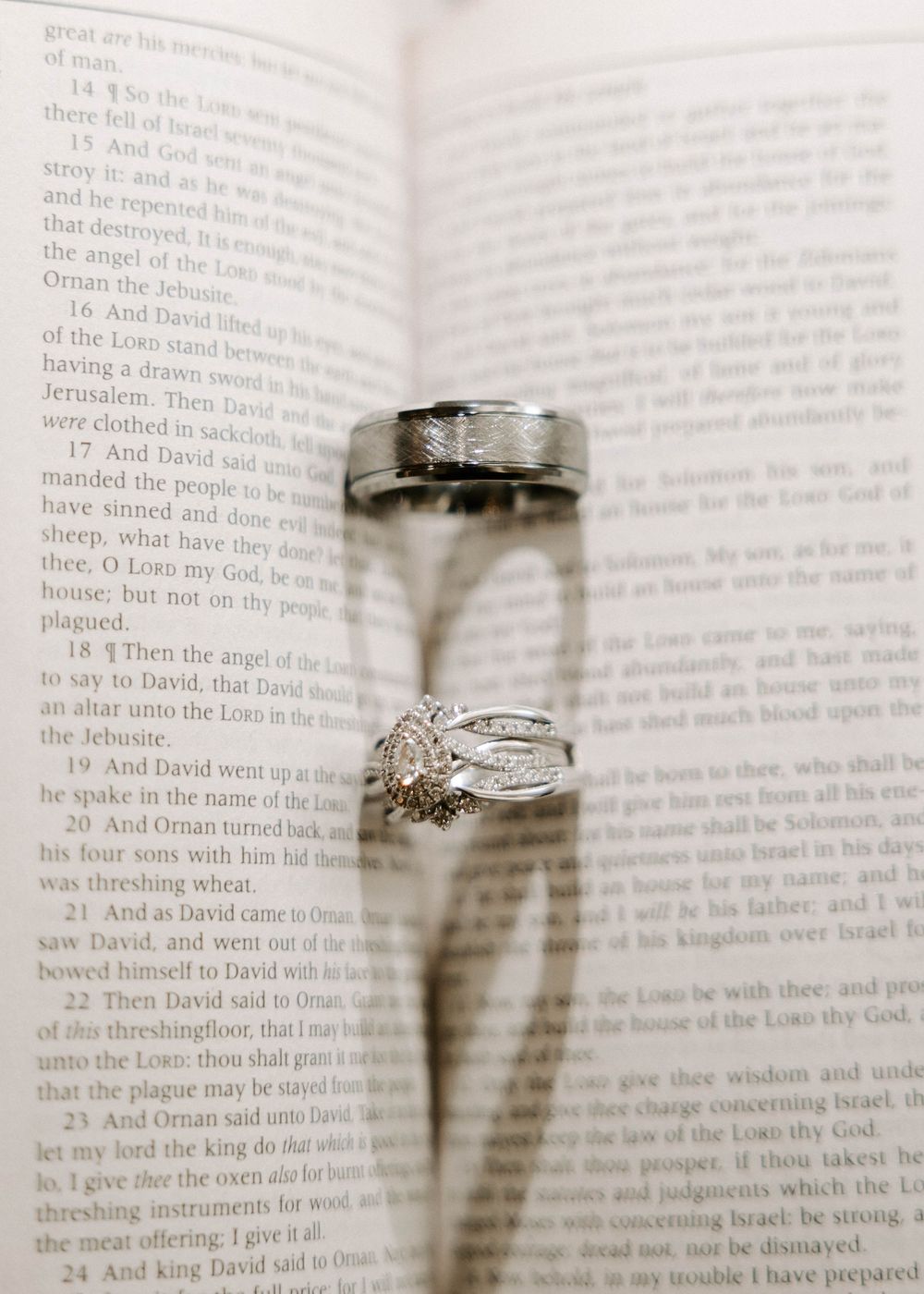 Wedding rings resting on an open Bible page, casting heart-shaped shadow, featuring diamond engagement ring and silver band.