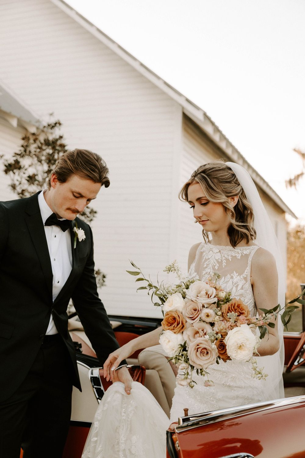 Bride and groom share romantic moments near vintage car during sunset wedding photoshoot.