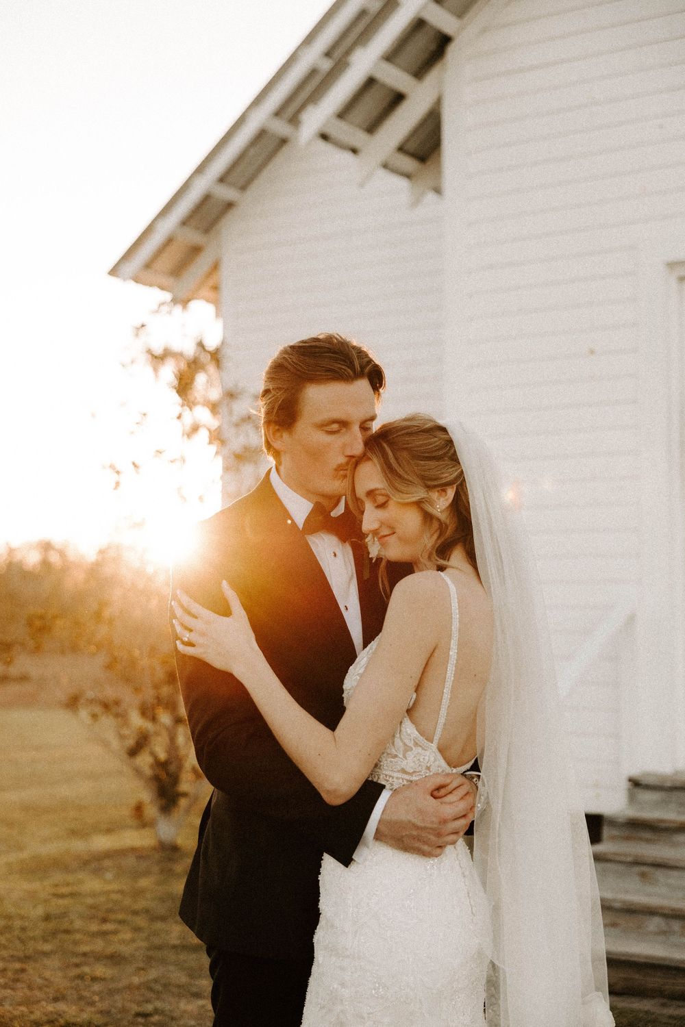 Romantic wedding couple embraces during golden hour sunset outside white chapel.