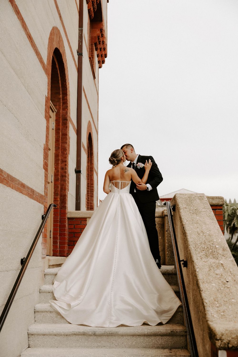 Bride and groom share a kiss on stone steps outside a brick building, her white ballgown train cascading down.