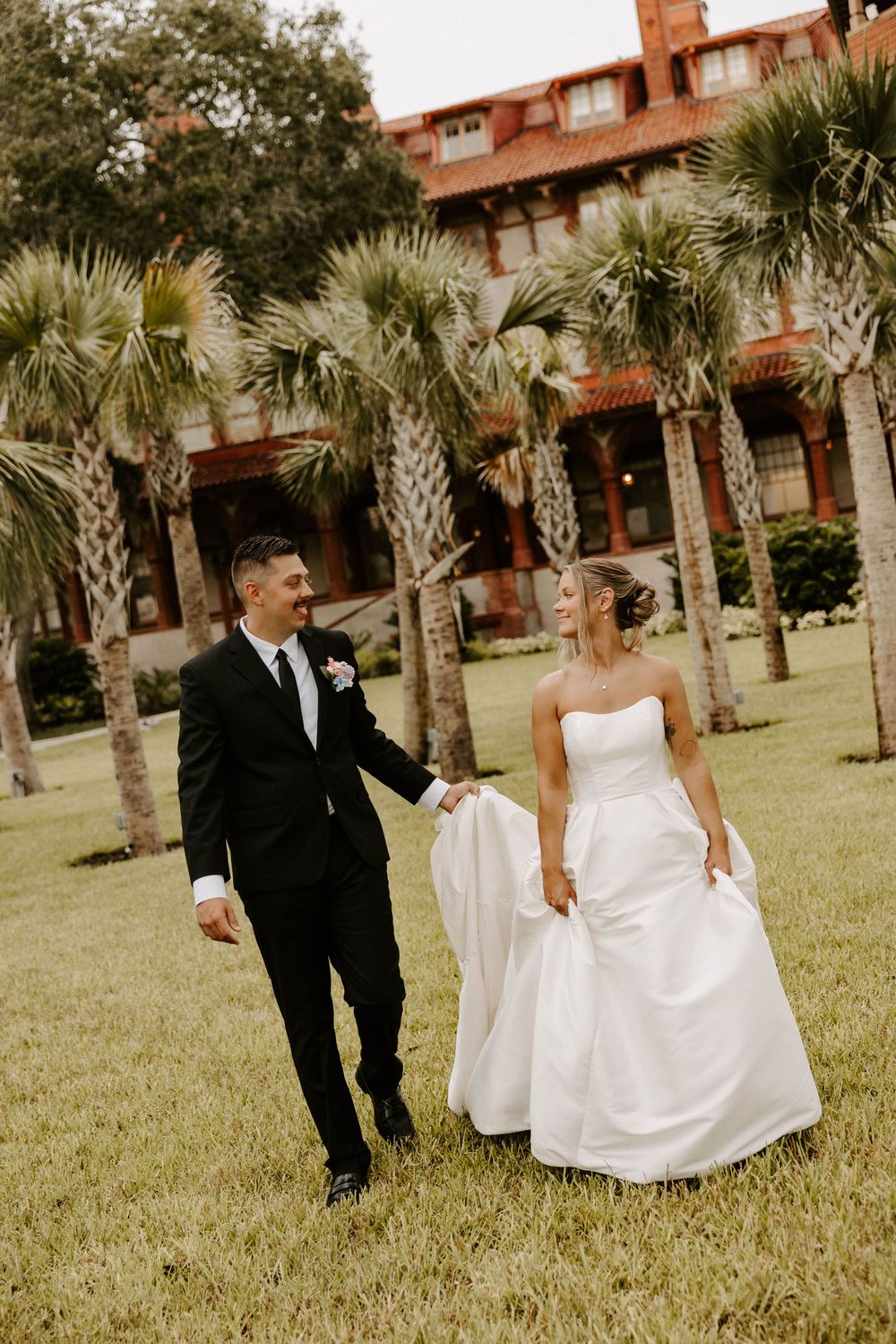Bride and groom walk hand in hand on green lawn with palm trees and historic red building backdrop.