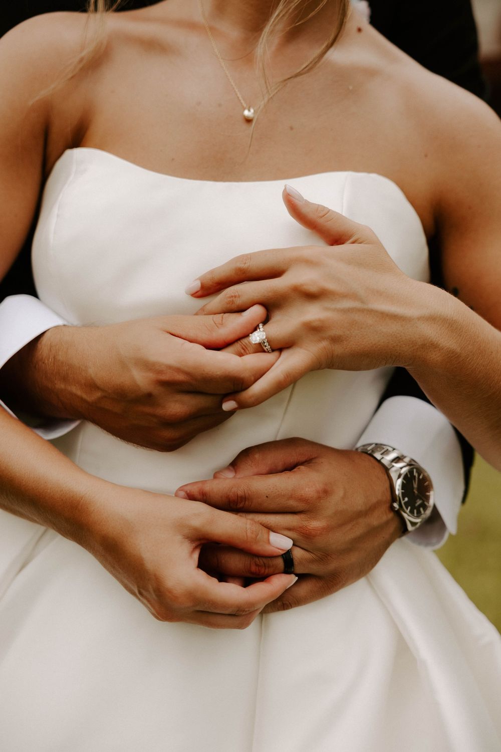 Bride and groom showing wedding rings, groom embracing bride in white strapless gown, wearing a Rolex watch.