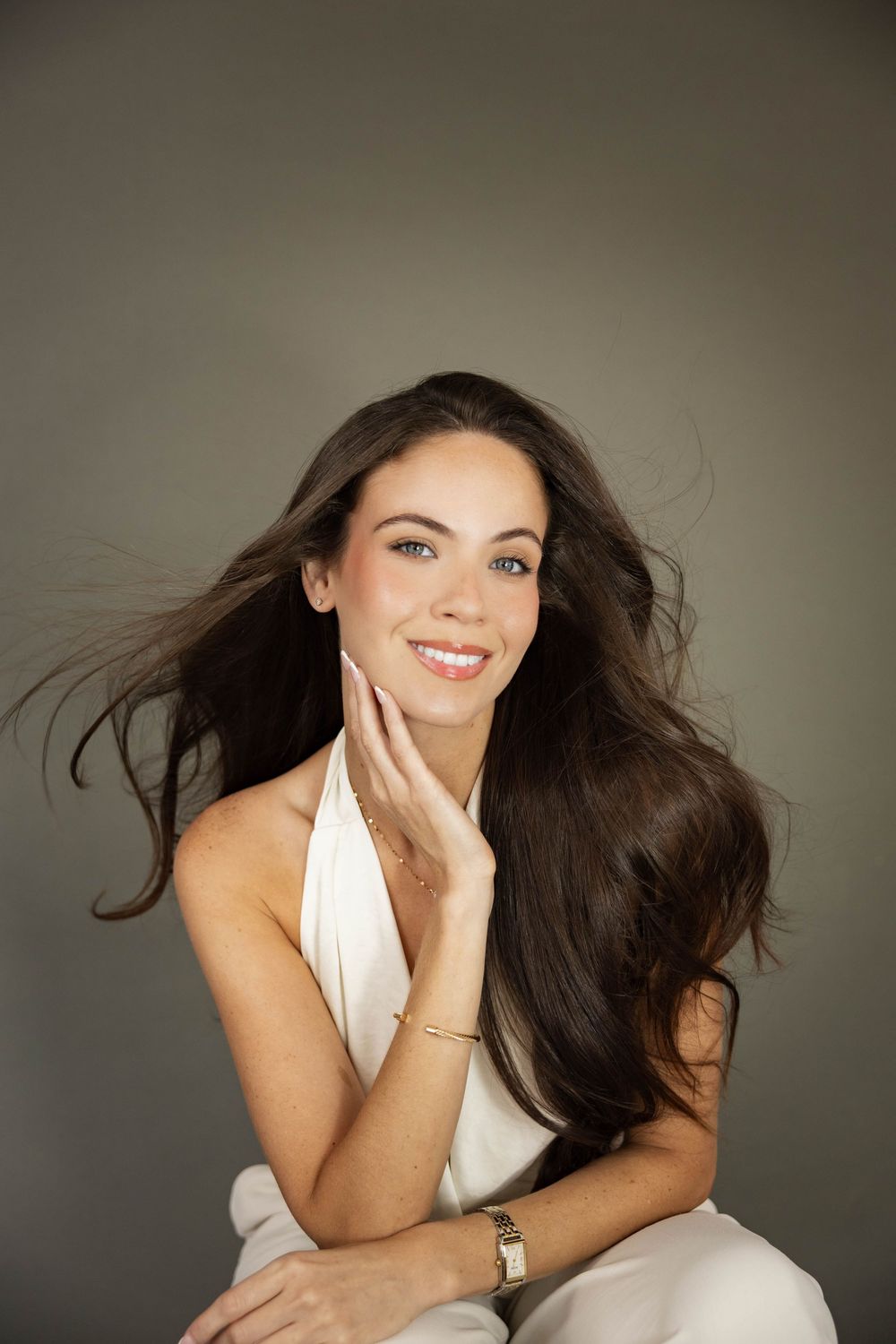Smiling brunette woman with flowing hair in white dress touching her face against gray background.