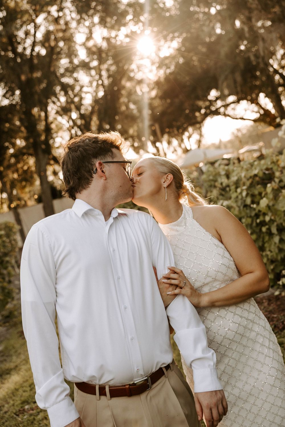 Couple sharing a romantic kiss outdoors at golden hour, man in white shirt and woman in white dress surrounded by trees.
