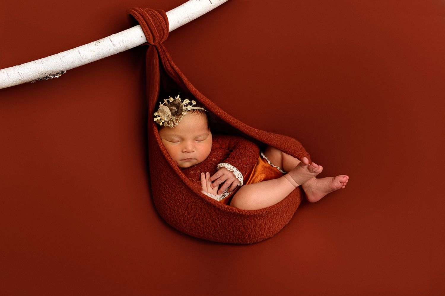 A sleeping baby wearing a gold crown lies peacefully in a rust-colored fabric hammock against a matching background.