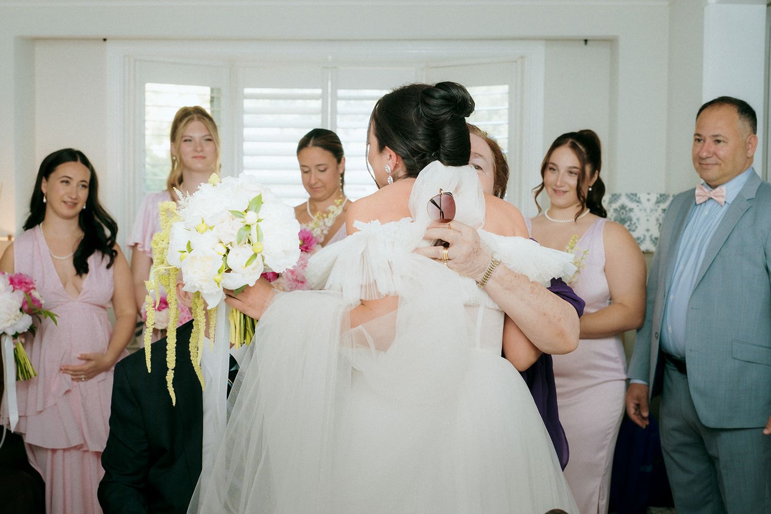 Group photo of wedding party in formal wear gathered in bright interior space.