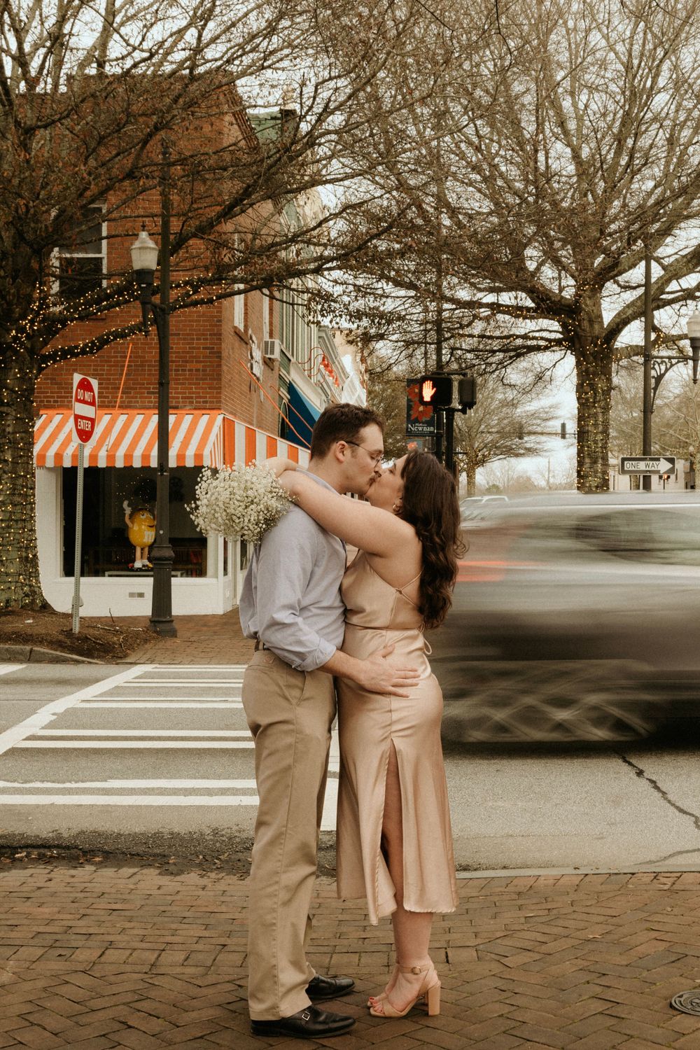 A couple shares a romantic embrace on a brick-paved street corner in formal attire.