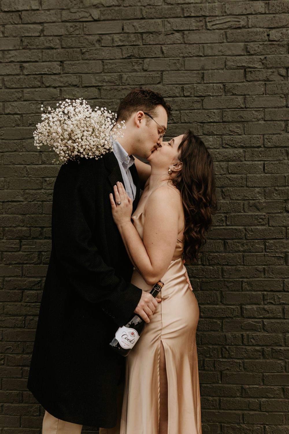 A formal couple embraces against a dark brick wall with baby's breath flowers.