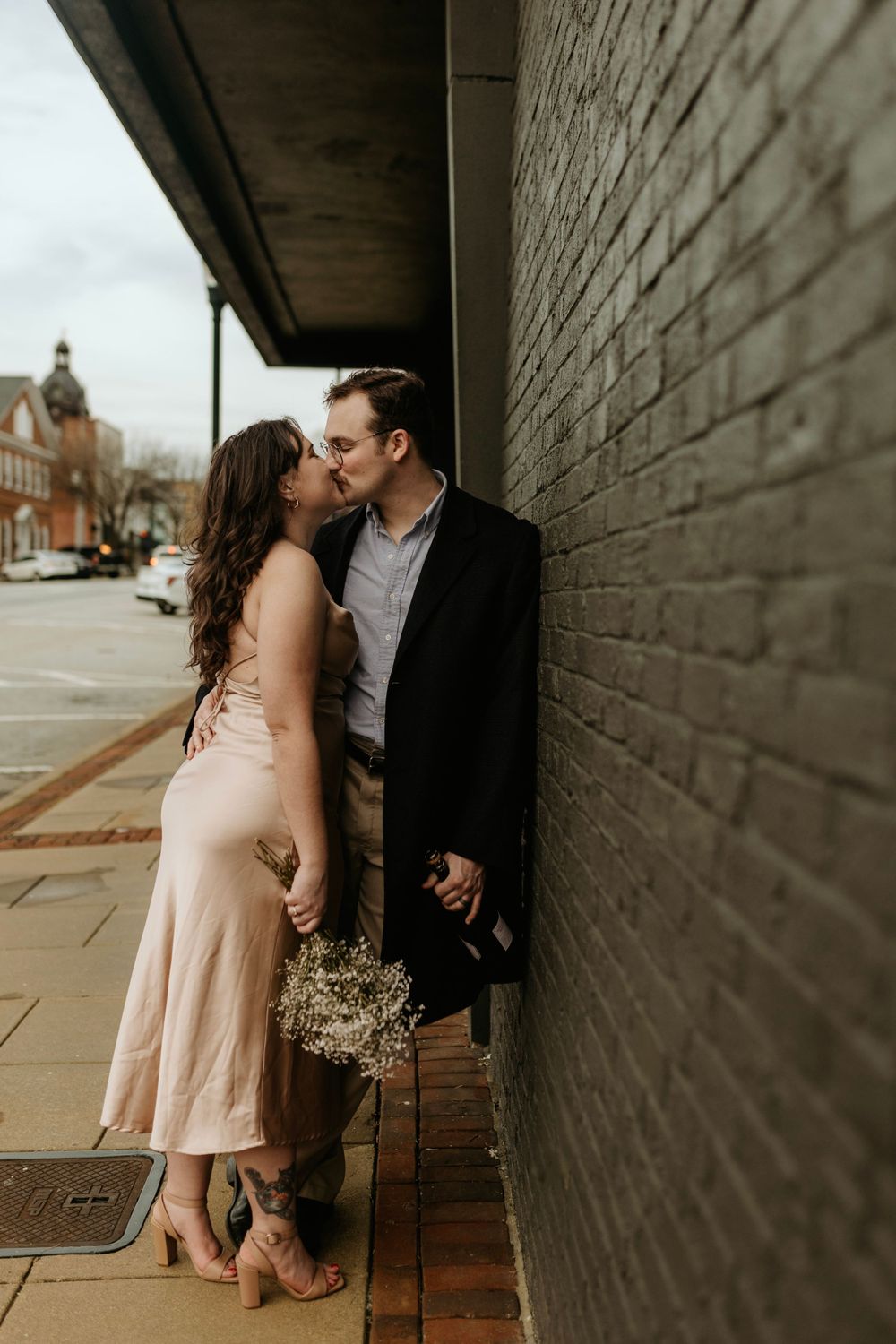 A romantic couple shares an intimate moment against a brick wall in their formal attire.