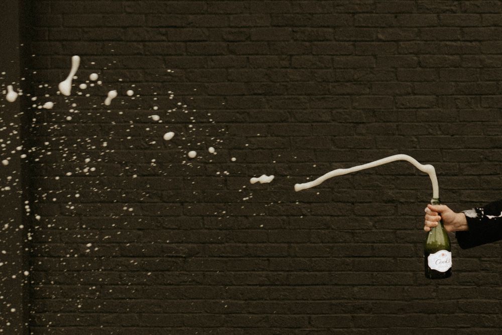 Champagne bottle popping with spray against dark brick wall backdrop.