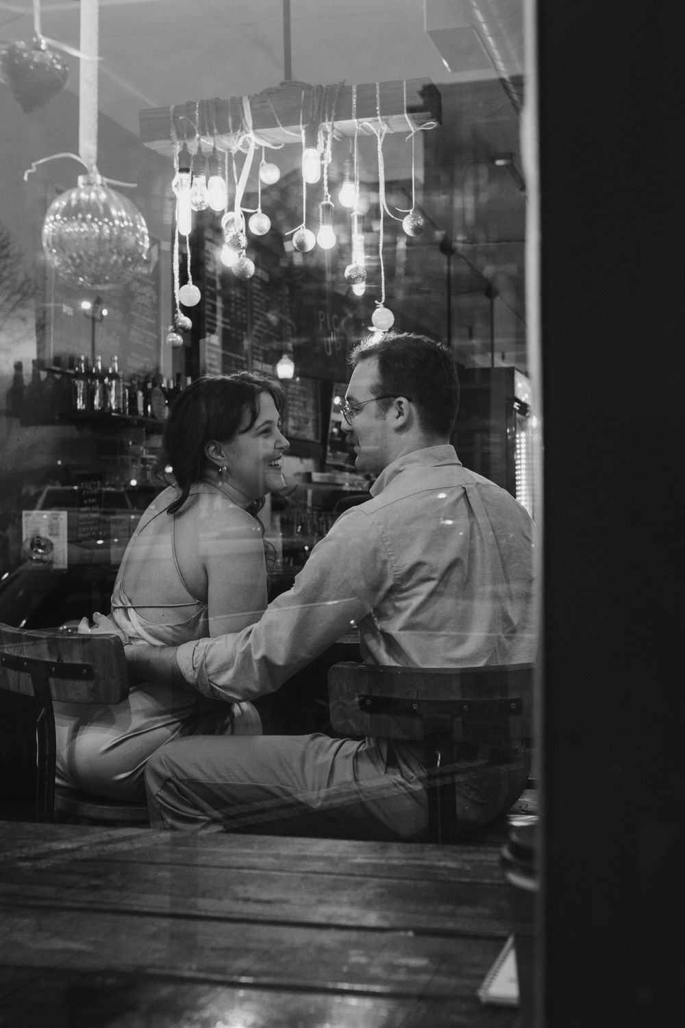 Black and white photo of a couple sharing an intimate moment in a restaurant with hanging lights.