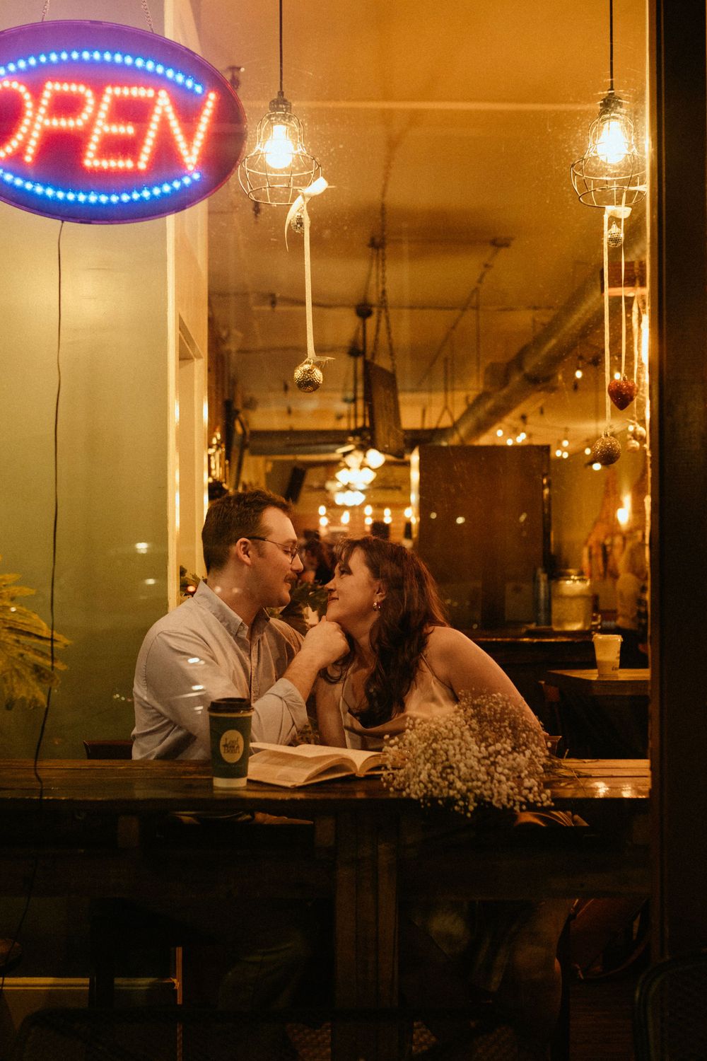 A couple shares a romantic moment in a cafe window with a neon open sign glowing.