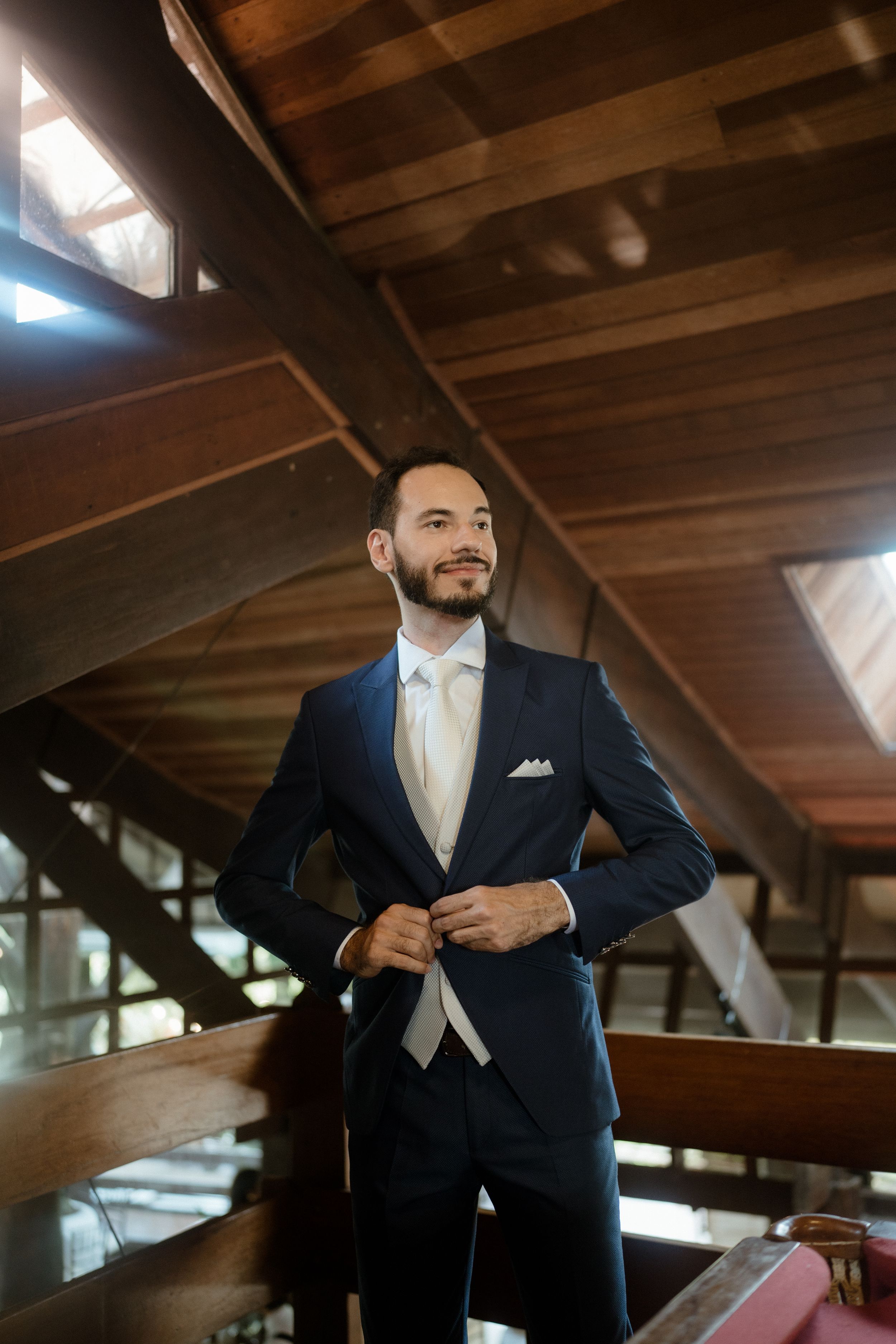 A person in a navy suit and cream tie adjusts their jacket in warm lighting.
