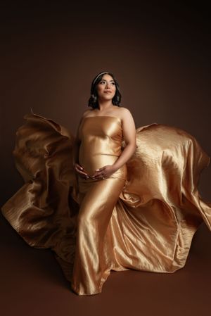 Dramatic maternity portrait with flowing gold fabric against dark brown background.