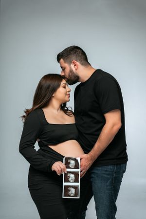 Expectant couple in black clothing sharing intimate moment while holding ultrasound photos.