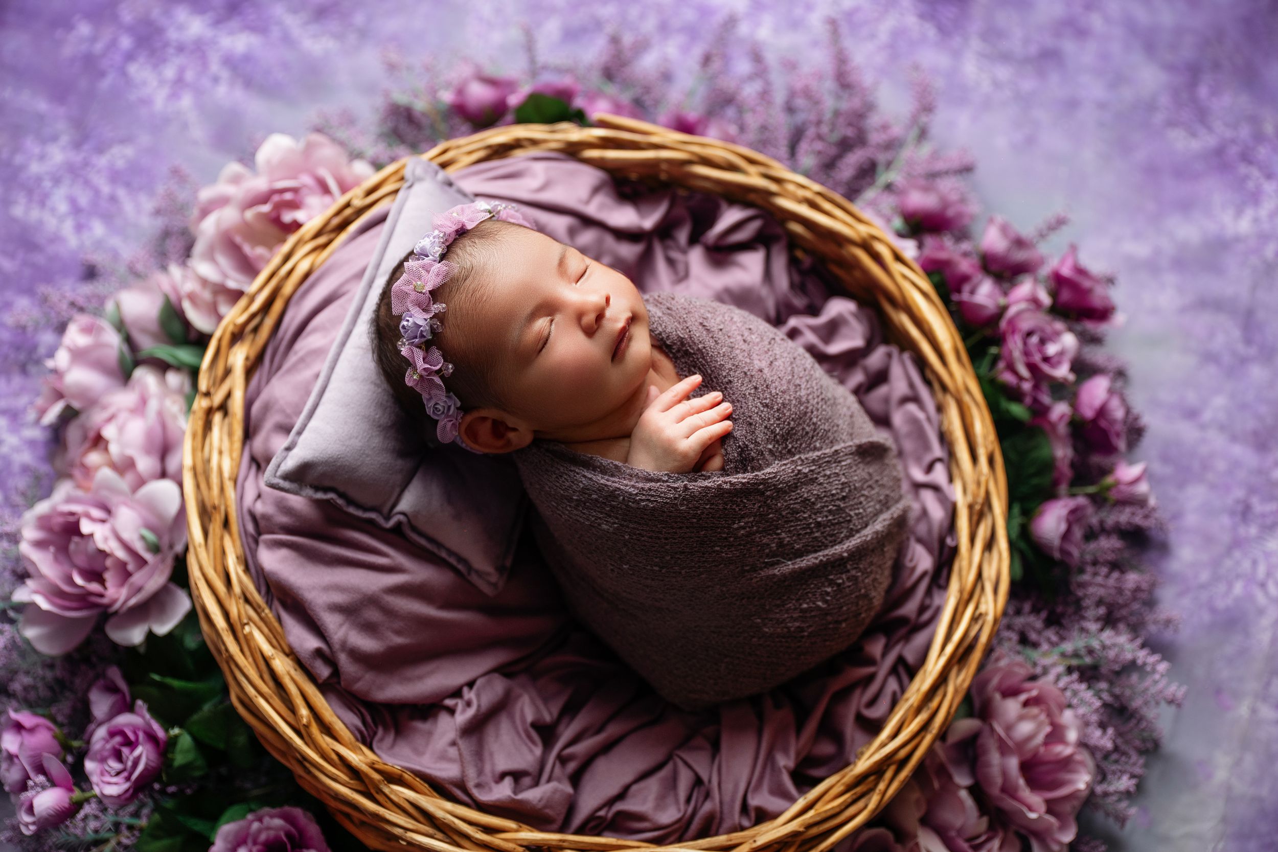 Newborn baby sleeping peacefully in a wicker basket wrapped in purple, surrounded by pink and lavender flowers.
