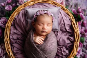 Newborn baby swaddled in purple wrap sleeping in wicker basket with floral headband and pink roses.