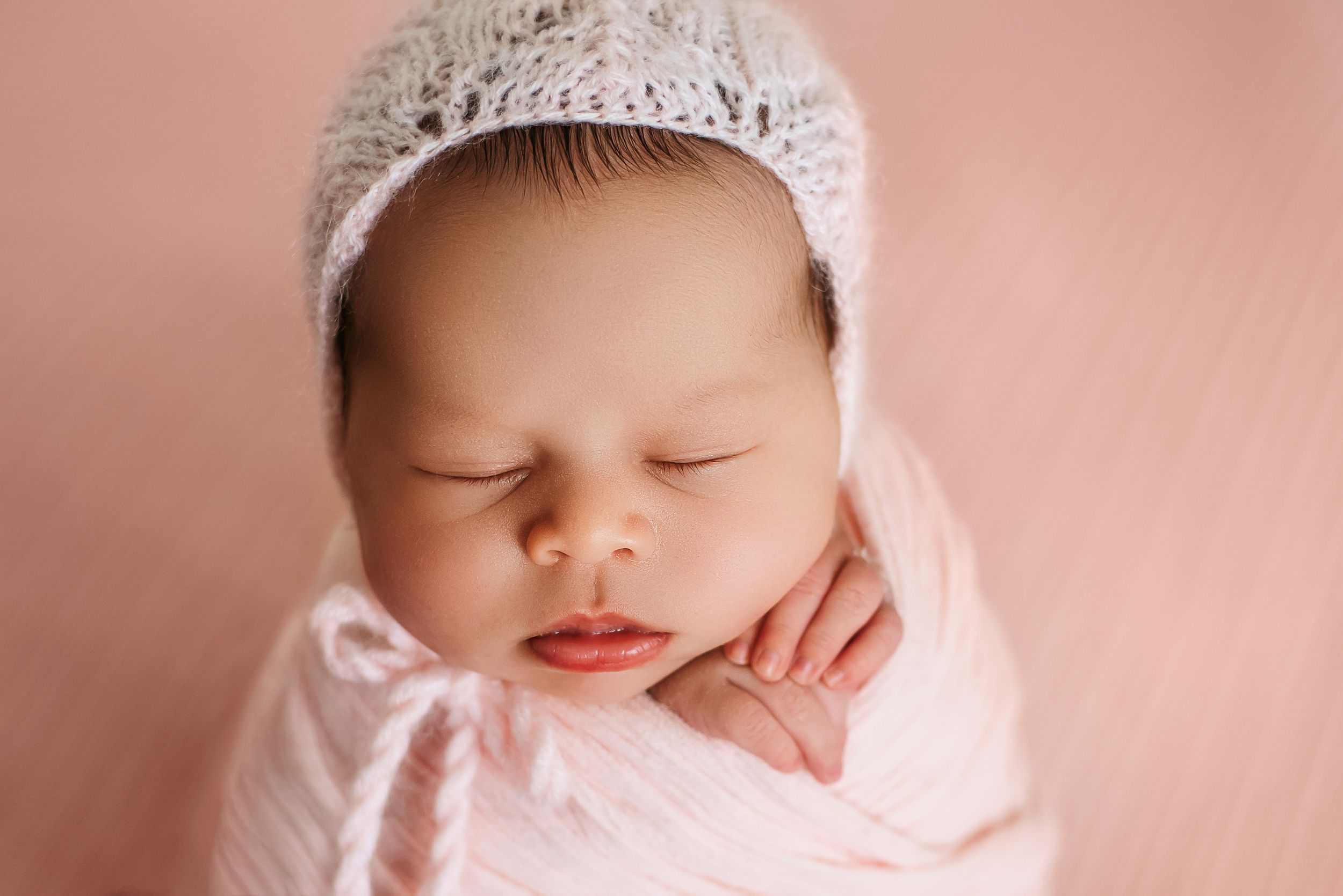 Peaceful newborn baby wearing knit bonnet sleeping on pink background.