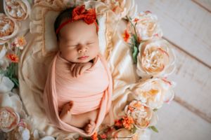 Sleeping newborn wrapped in peach swaddle surrounded by peachy roses on white wooden floor.