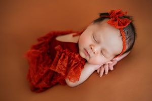 Sleeping newborn in red lace dress with matching bow headband on brown backdrop.