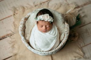 Newborn baby nestled in white round basket wearing floral crown and swaddled in white fabric.