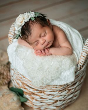 Sleeping newborn baby wrapped in white cloth lying in wicker basket with delicate floral headpiece.