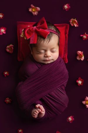 Sleeping baby wrapped in burgundy fabric with red bow on dark background surrounded by flowers.