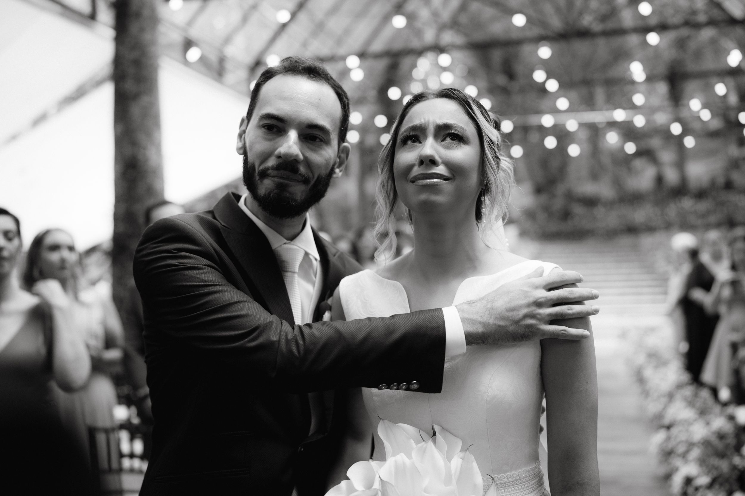 A tender moment captured during a wedding ceremony in a greenhouse setting with string lights.