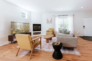Bright modern living room with hardwood floors, white walls, fireplace, mid-century chairs, and neutral sofa on patterned rug.