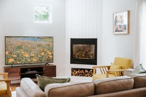 Cozy modern living room with large TV, fireplace, yellow armchair, and neutral sofa in a bright white interior.