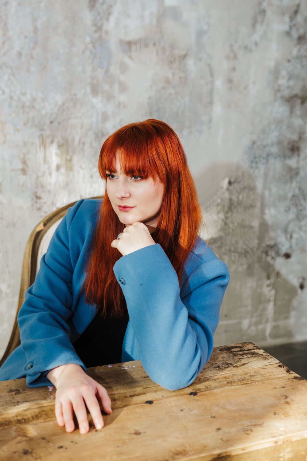 A vibrant redhead wearing a blue sweater sits pensively at a rustic wooden table against a textured wall.