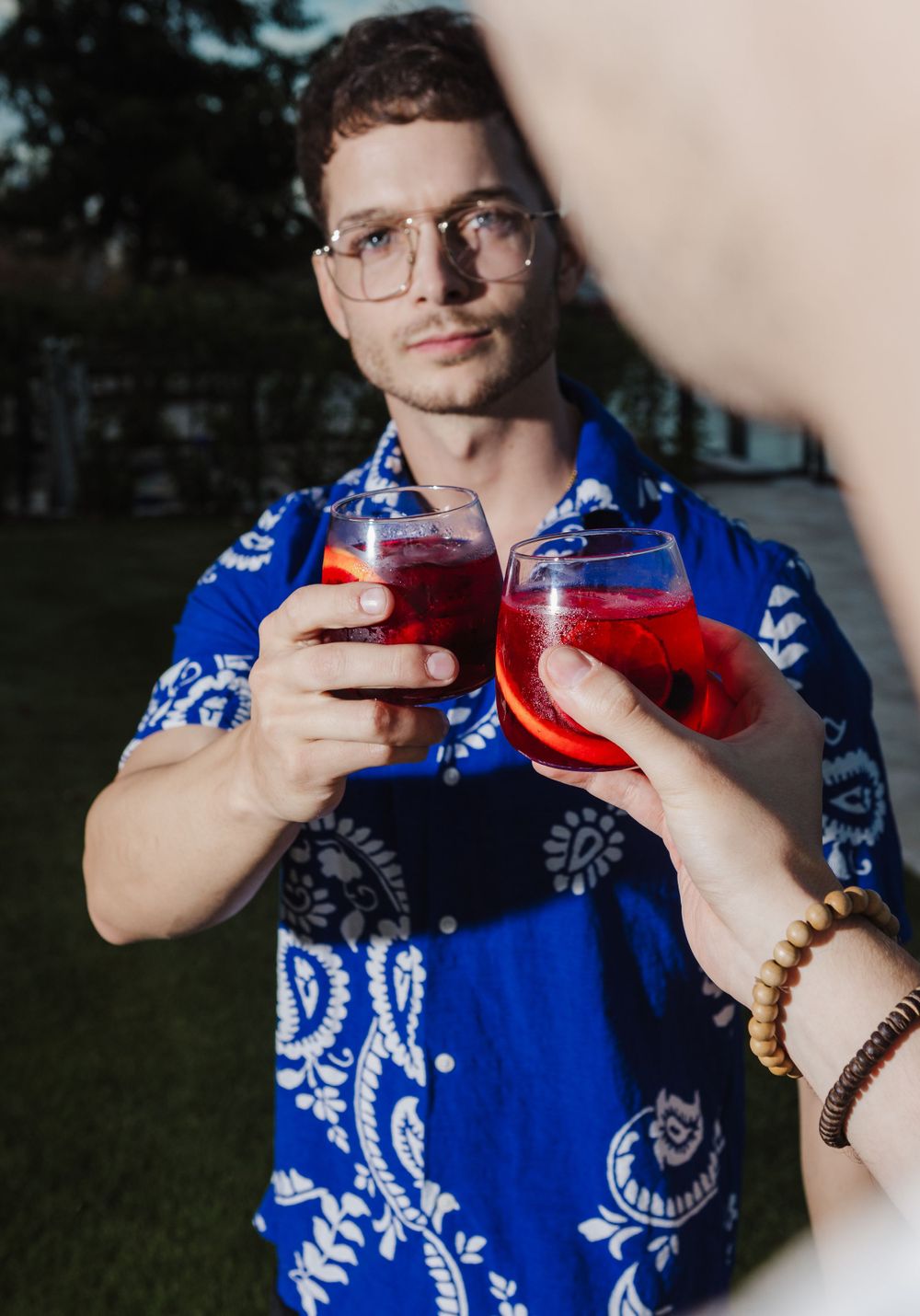 Person in blue patterned shirt holding red cocktails in outdoor setting at dusk.