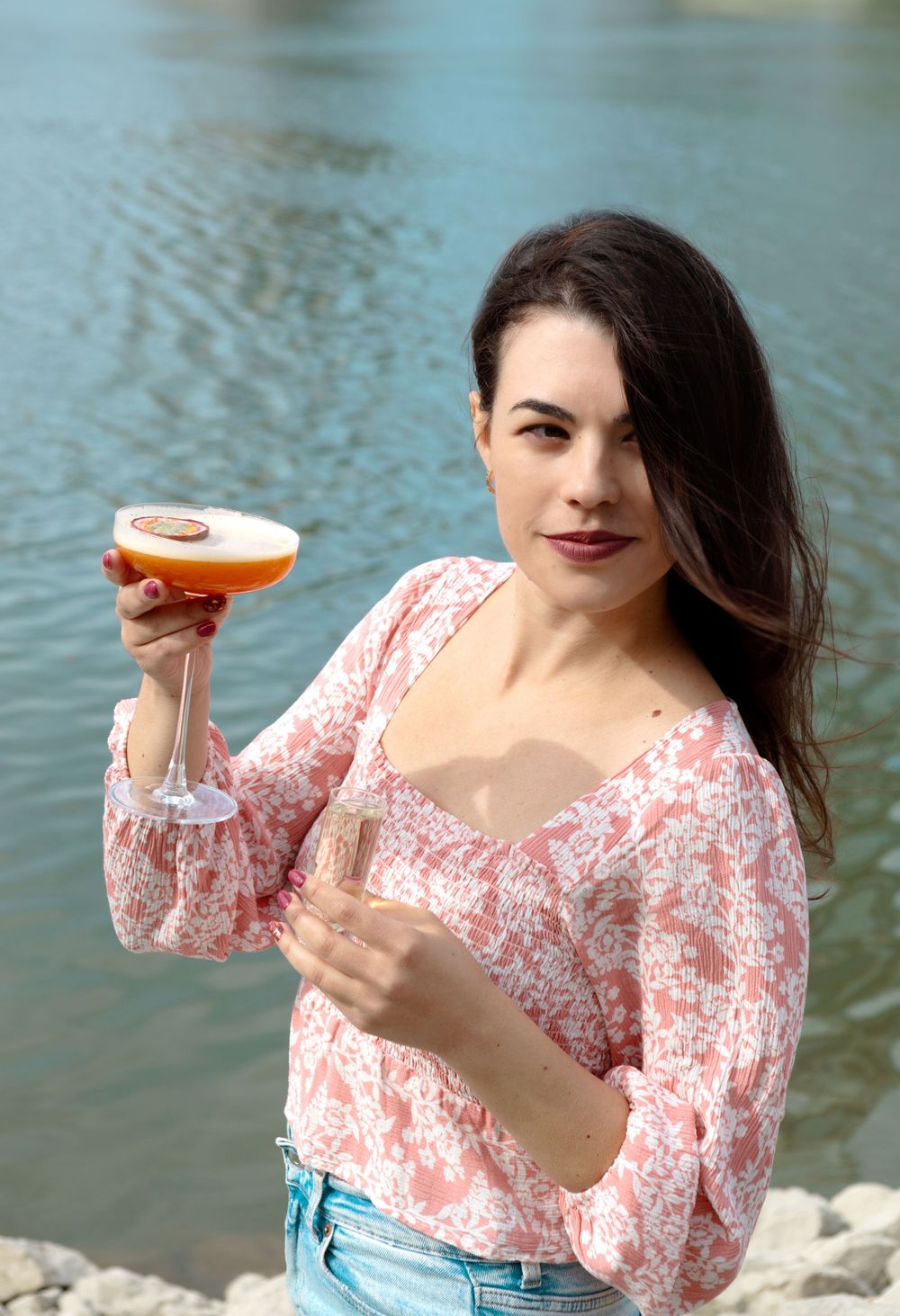 Woman in floral pink top holding orange cocktail by waterfront.