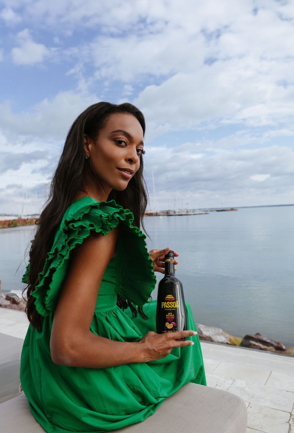 Woman in emerald green dress poses with bottle by waterfront marina.
