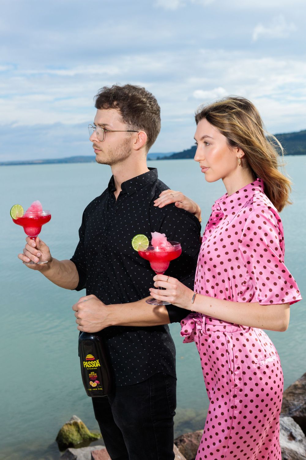 Couple holding red cocktails by the waterfront in polka dot dress and black attire.