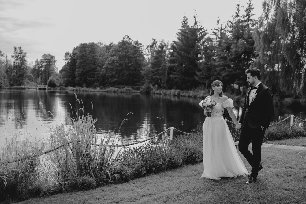 A black and white wedding portrait beside a tranquil lake surrounded by trees.