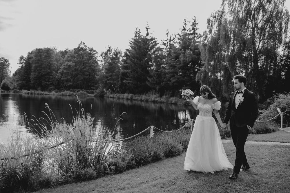A black and white wedding photo shows a couple walking along a serene lakeside path surrounded by evergreen trees.