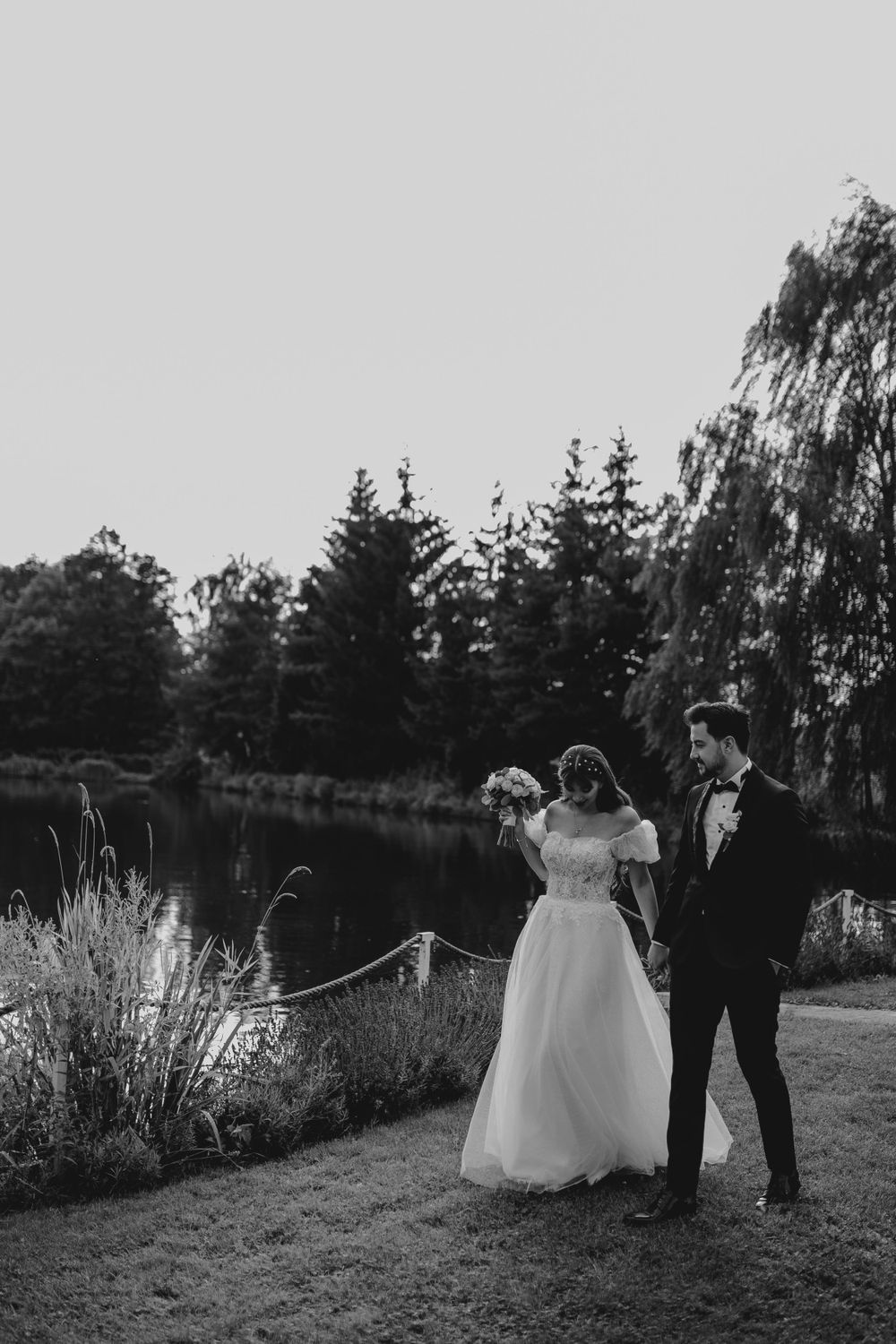 A romantic wedding couple poses by a tranquil pond with drooping willow trees and evergreens in the background.