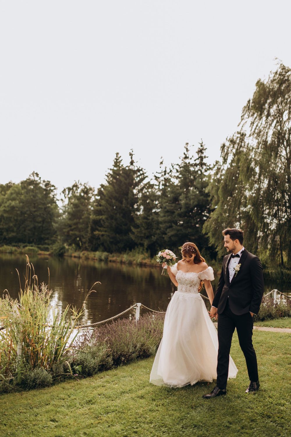 A wedding couple shares a tender moment beside a peaceful lake while surrounded by tall pine trees.