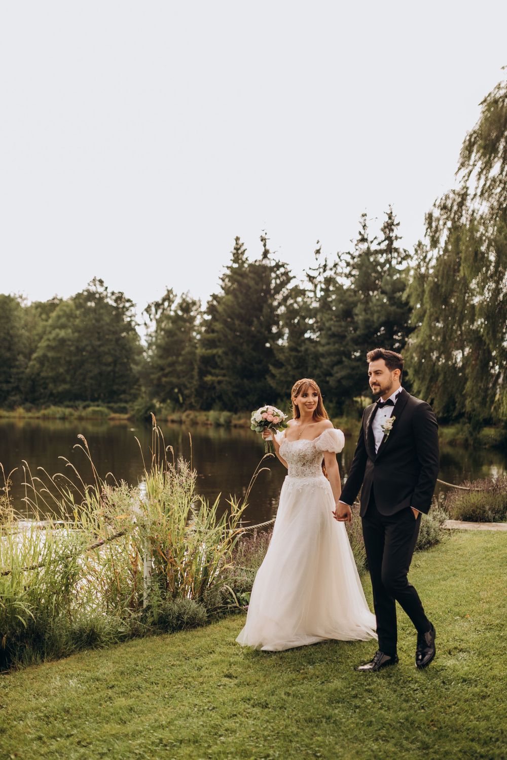 A couple in wedding attire walks along a landscaped path beside a serene pond with pine trees in the background.