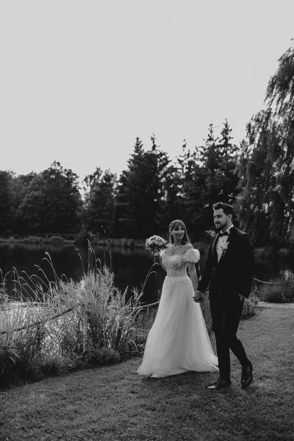 A monochrome wedding photo captures a couple strolling along a lakeside path with tall grass and trees.